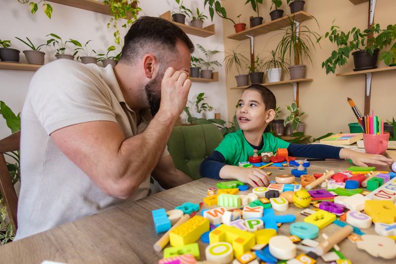 A man and child sit at a table filled with colourful wooden letters and toys. They playfully interact in a room adorned with potted plants.